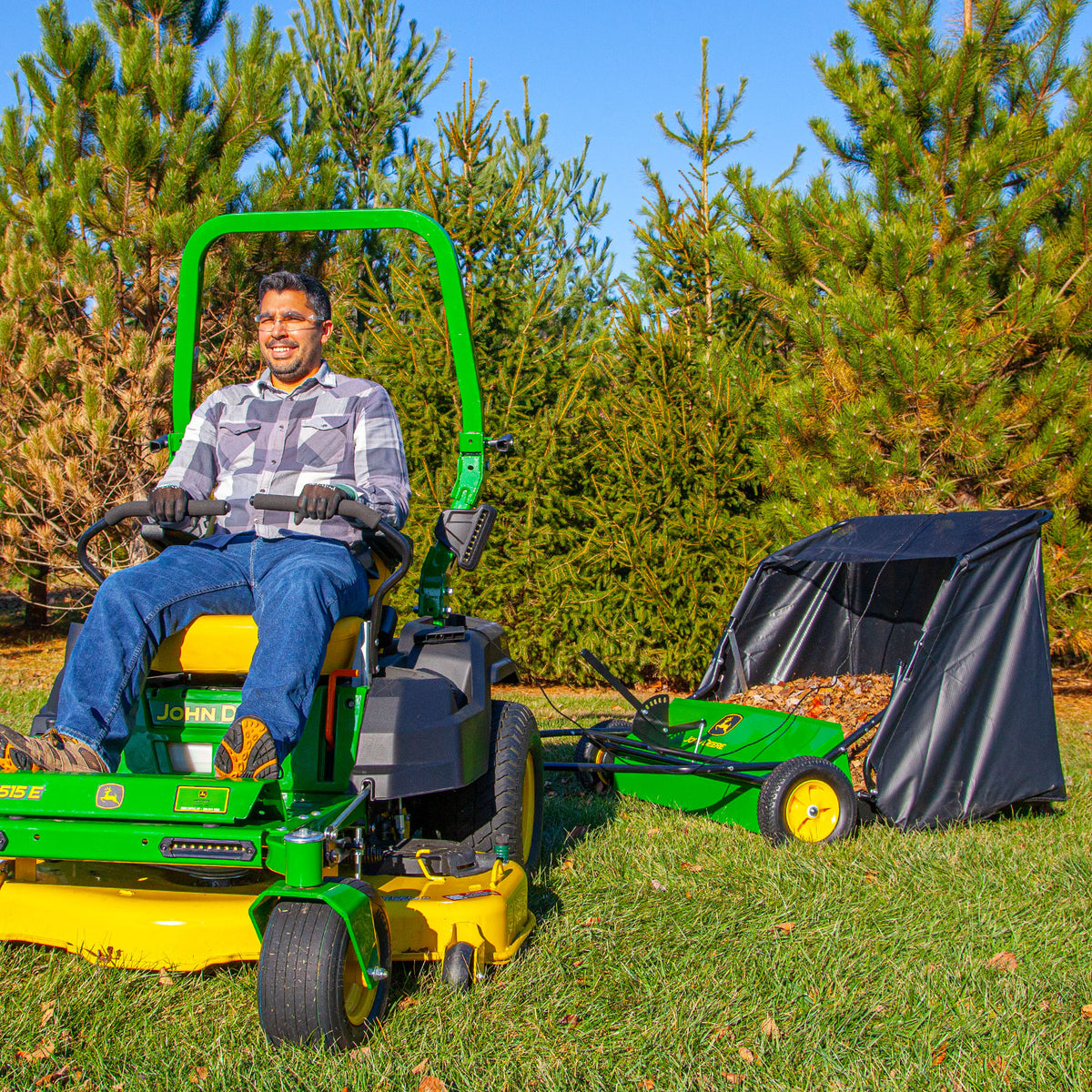 Wearing a plaid shirt and sunglasses, a person maneuvers a green John Deere ride-on lawnmower with a 42 in. path, towing a black and green 24 cu. ft. John Deere Tow-Behind Lawn Sweeper, effortlessly making yard work cool as they navigate through grass bordered by pine trees.