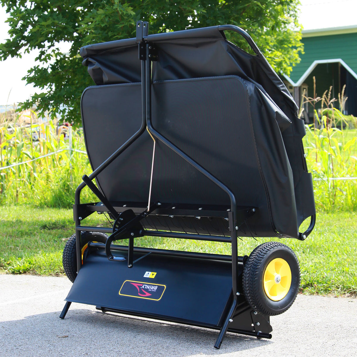The Brinly Parts 42″ Lawn Sweeper with Dethatcher (STS-42BHDK-A) in black, featuring yellow wheels, stands upright on a driveway with green grass, corn stalks, and a green building visible in the background.