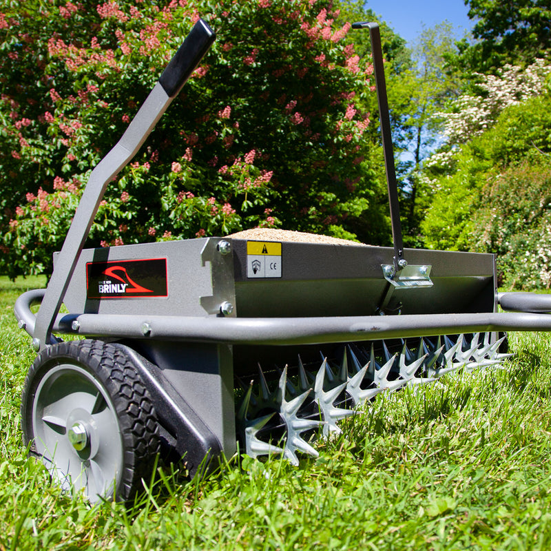 The Brinly Parts 40 Combination Aerator Spreader in Hammered Gunmetal (AS2-40BH-S) features spiked wheels and a metal frame with a convenient handle, set on grassy terrain. In the background, lush bushes with green leaves and red flowers flourish under the clear blue sky.