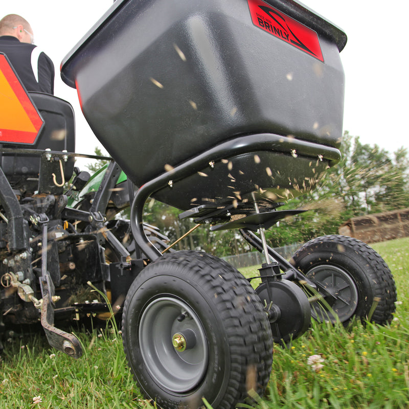 A close-up shows a Brinly Parts 175 Lb. Tow-Behind Spreader (BS36BH) attached to a vehicle dispersing seeds or fertilizer on a grassy field. The rust-proof polyethylene spreader, with large wheels, is in action as someone in a black jacket drives, with trees visible in the background.