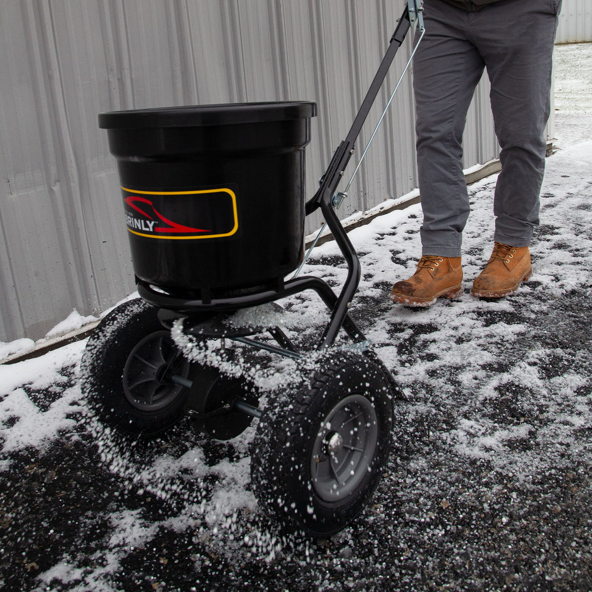 A person in brown boots pushes a Brinly Parts 50 LB. Push Spreader with Side Deflector & Hopper Cover (P20-500BHDF-A) across a snowy, icy surface beside a corrugated metal wall.