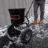 A person in brown boots pushes a Brinly Parts 50 LB. Push Spreader with Side Deflector & Hopper Cover (P20-500BHDF-A) across a snowy, icy surface beside a corrugated metal wall.