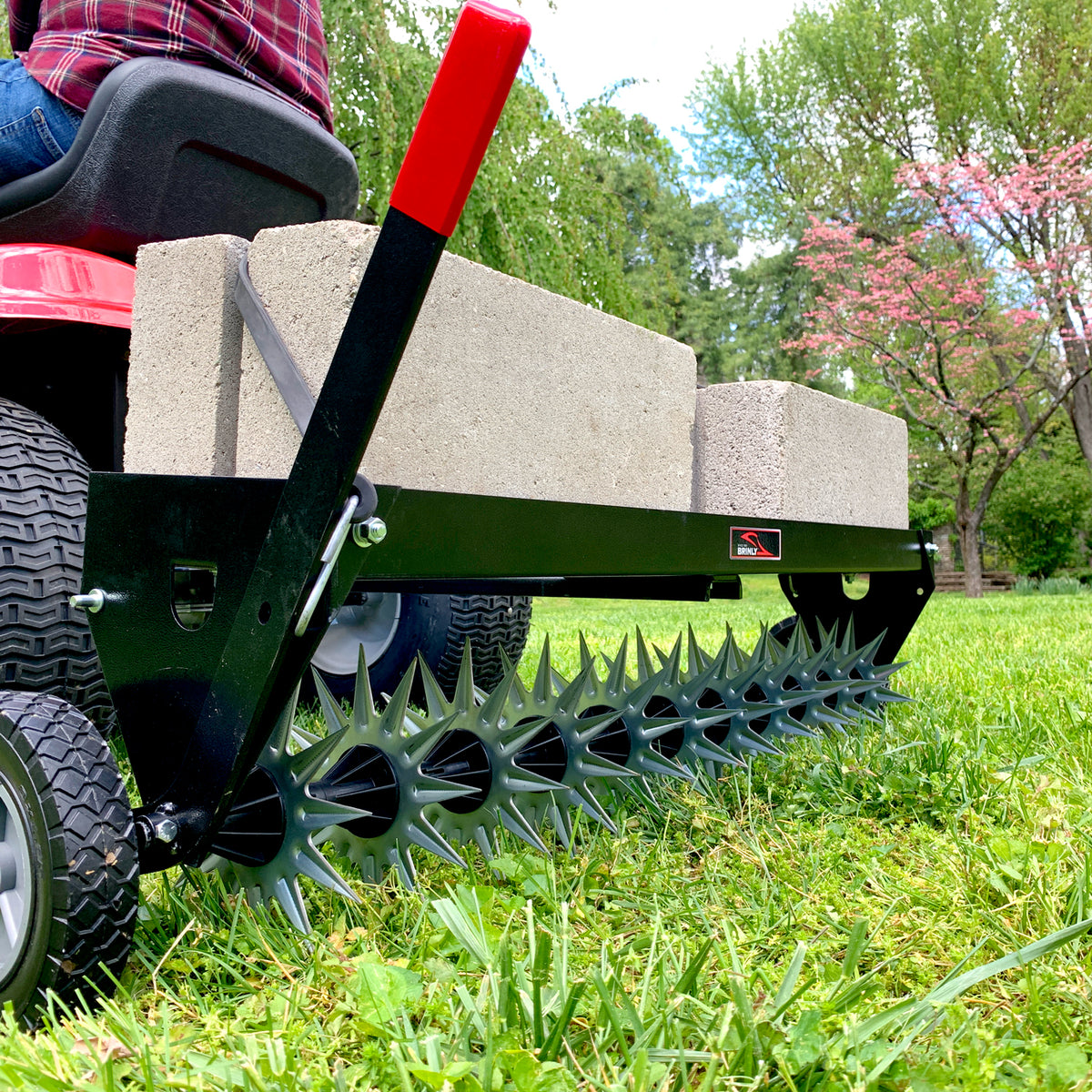 A Brinly Parts 40″ Tow-Behind Spike Aerator (SAT2-40BH-G) is hitched to a red lawn tractor. It features spiked wheels and three concrete block weights, effectively aerating a grassy lawn framed by vibrant green trees and a blossoming pink tree in the background.