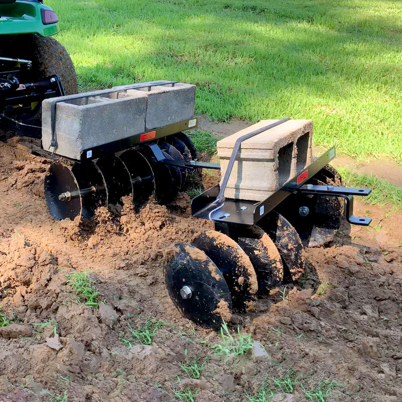 Brinly Parts Sleeve Hitch Disc Harrow | DD-551BH, featuring black metal discs, is attached to a tractor for efficient soil prep. Added concrete blocks enhance effectiveness. Vibrant green grass and freshly churned soil hint at successful cultivation ahead.