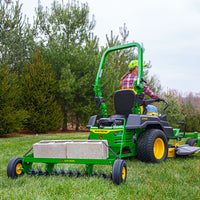 A person in a plaid shirt and cap operates a John Deere lawn mower with a 40 Spike Aerator attachment, featuring steel tine stars and a Rigid Steel Weight Tray loaded with concrete blocks, mowing grass bordered by pine trees.