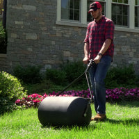 A person in a red plaid shirt and cap is using the Brinly Parts 28 Gallon Push or Tow Poly Lawn Roller (PRC-242BH) on a grassy area in front of a stone building with windows, with colorful flowers and bushes adding a vibrant backdrop.