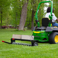 A person drives a green lawn tractor, towing the Brinly Parts 40″ Spike Aerator with Transport Wheels & 3D Galvanized Steel Tines (SAT-401BH) loaded with concrete blocks across a grassy yard for effective aeration.