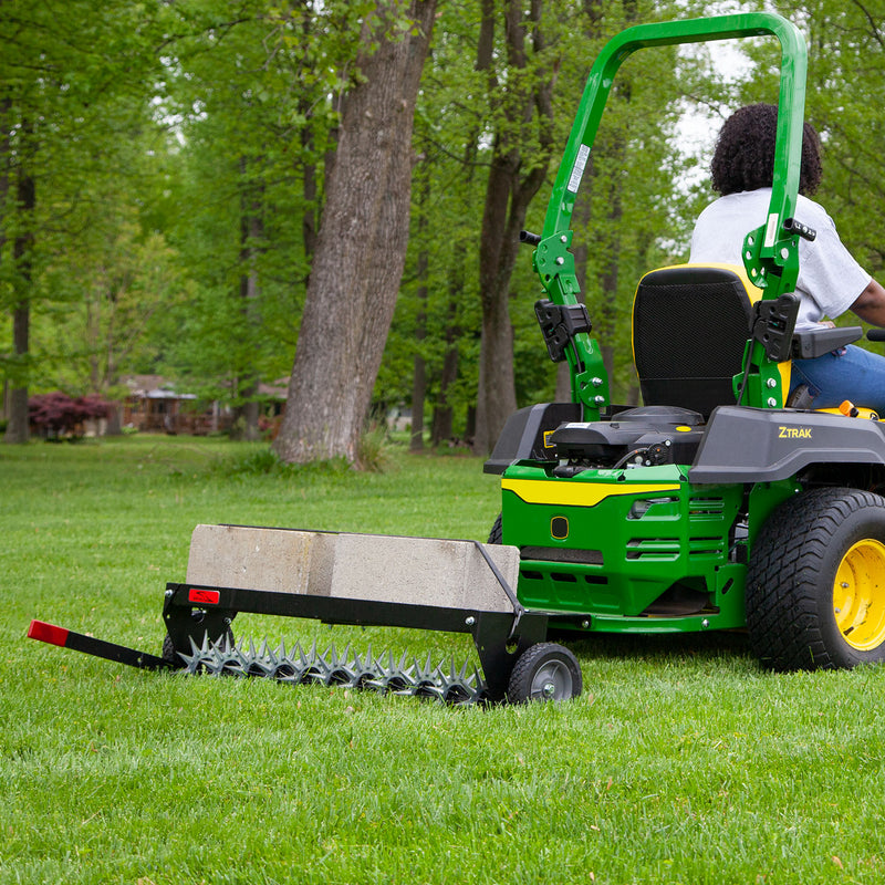 A person drives a green lawn tractor, towing the Brinly Parts 40″ Spike Aerator with Transport Wheels & 3D Galvanized Steel Tines (SAT-401BH) loaded with concrete blocks across a grassy yard for effective aeration.