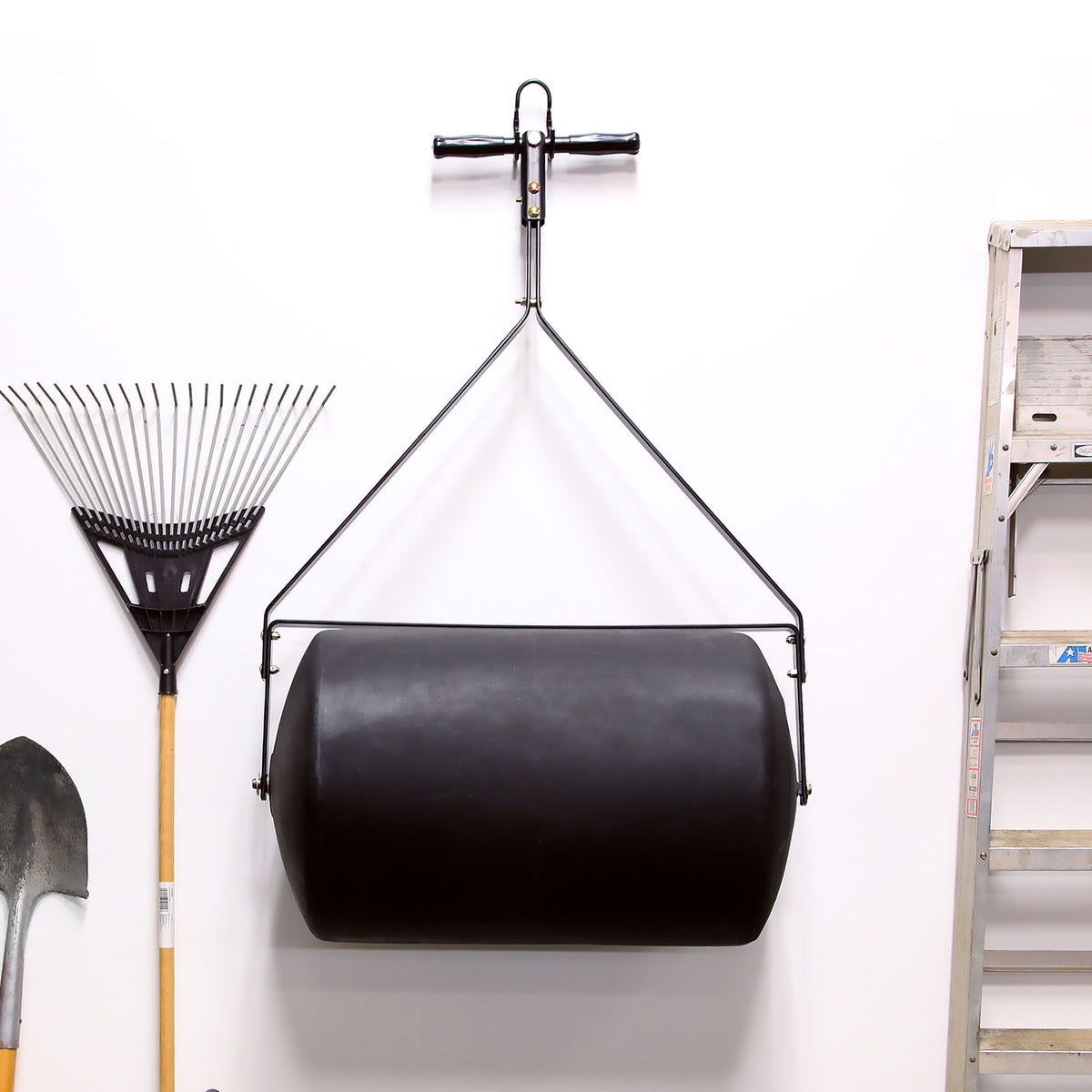 A 28 Gallon Push or Tow Poly Lawn Roller by Brinly Parts hangs using a pulley system. To the left, a metal rake and shovel rest against the white wall, while part of a metal ladder is visible to the right.