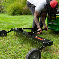 A person in a red cap and gray shirt adjusts a Brinly Parts 48 Tow-Behind Dethatcher (DT-480BH) hitched to a green lawn tractor on grassy terrain, tending to essential lawn care with trees and bushes in the backdrop.