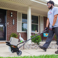 Wearing safety gear, a man uses a Brinly Parts 20 Push Spike Aerator with 3D Galvanized Steel Tines and a concrete block to perforate soil near the lawns edge in front of a brick house with a porch.