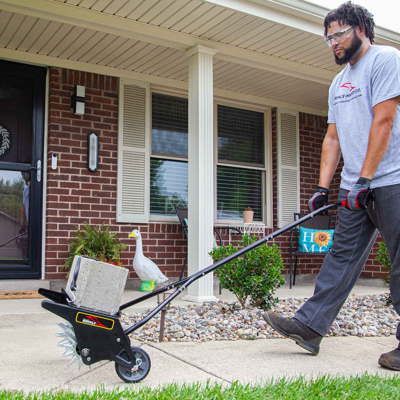 Wearing safety gear, a man uses a Brinly Parts 20 Push Spike Aerator with 3D Galvanized Steel Tines and a concrete block to perforate soil near the lawns edge in front of a brick house with a porch.