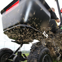 A close-up of a riding lawn mower demonstrates the Brinly Parts 175 Lb. Tow-Behind Spreader | BS36BH dispersing grass seeds or fertilizer. The rust-proof polyethylene container distributes seeds while the wheels glide over lush, green grass.