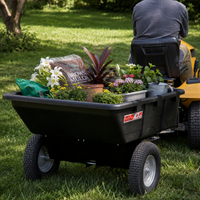 A person drives a lawn tractor pulling the Brinly Parts 10 Cu. Ft. Poly Cart (Model PCT-101BH) loaded with flowers, potted plants, gardening supplies, and a bag of brown mulch across a green grassy yard.