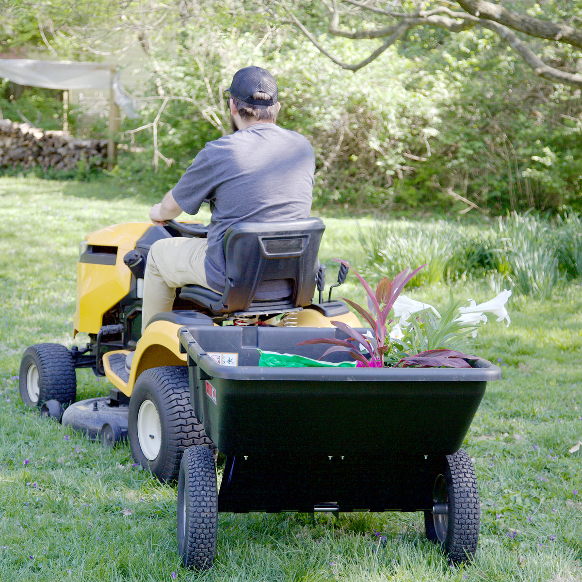 A person in a cap and gray shirt drives a yellow riding mower with a Brinly Parts 10 Cu. Ft. Poly Cart (PCT-101BH) attached, hauling plants and gardening supplies across a grassy yard.
