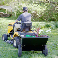 A person in a cap and gray shirt drives a yellow riding mower with a Brinly Parts 10 Cu. Ft. Poly Cart (PCT-101BH) attached, hauling plants and gardening supplies across a grassy yard.