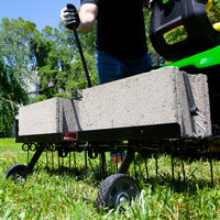 Wearing gloves, a person adjusts the handle on a Brinly Parts 40″ Tow-Behind Dethatcher DT2-40BH-G, weighted with concrete blocks on a lush lawn. As prongs and wheels assist in removing thatch, trees and a house create a picturesque backdrop.