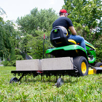 A man in a red cap and black shirt drives a green riding mower, efficiently dethatching the lawn using the Brinly Parts 40″ Tow-Behind Dethatcher | DT2-40BH-G, which is towing a metal frame weighted with concrete blocks. Trees and houses are visible in the background.