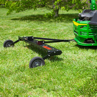 A black Brinly Parts 48” Tow-Behind Dethatcher (DT-480BH) is attached to a green ride-on mower on a grassy field. It features multiple black spikes and two wheels for lawn care. Bright green grass and trees are visible in the background.