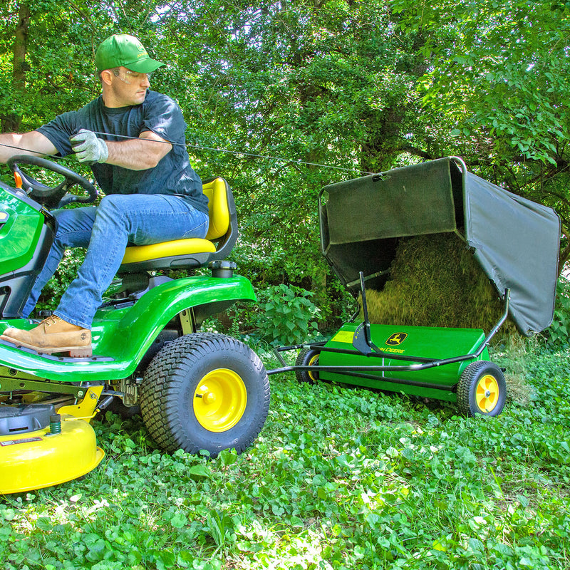 A person on a green and yellow John Deere lawn tractor tows a John Deere 42 in. 24 cu. ft. Tow-Behind Lawn Sweeper filled with grass in a lush green yard surrounded by trees, wearing a green cap, gloves, blue jeans, and tan shoes.