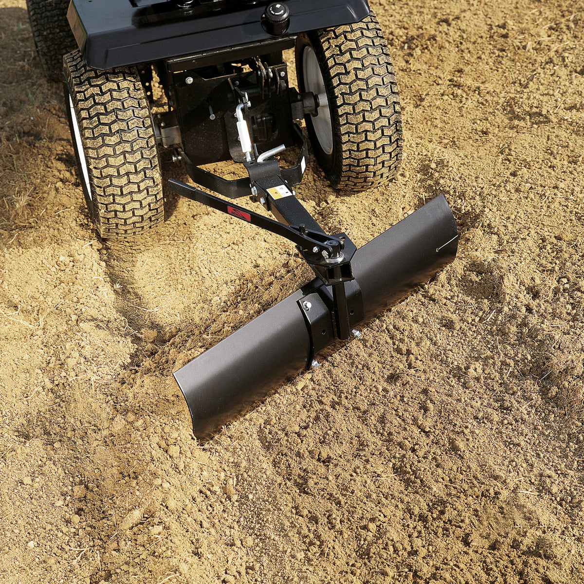 A close-up shows a Brinly Parts 42” Sleeve Hitch Rear Blade (BB-562) in action, seamlessly leveling soil with a tractor. The black rear blade is visible, along with the tractors large, textured tires amid loose dirt.