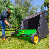 A person in a green cap adjusts the handle of a John Deere 42 Tow-Behind Lawn Sweeper (LPSTS-42JD / STS-42JD) with high-capacity hamper, set on a grassy lawn surrounded by trees and greenery on a sunny day.