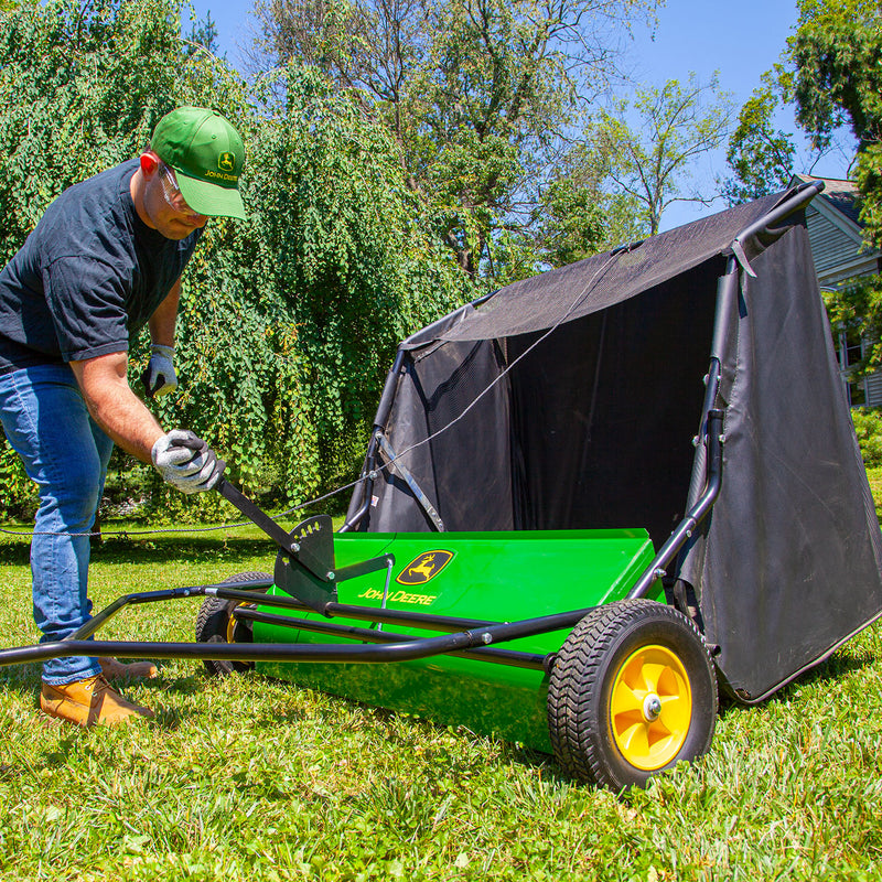 A person in a green cap adjusts the handle of a John Deere 42 Tow-Behind Lawn Sweeper (LPSTS-42JD / STS-42JD) with high-capacity hamper, set on a grassy lawn surrounded by trees and greenery on a sunny day.