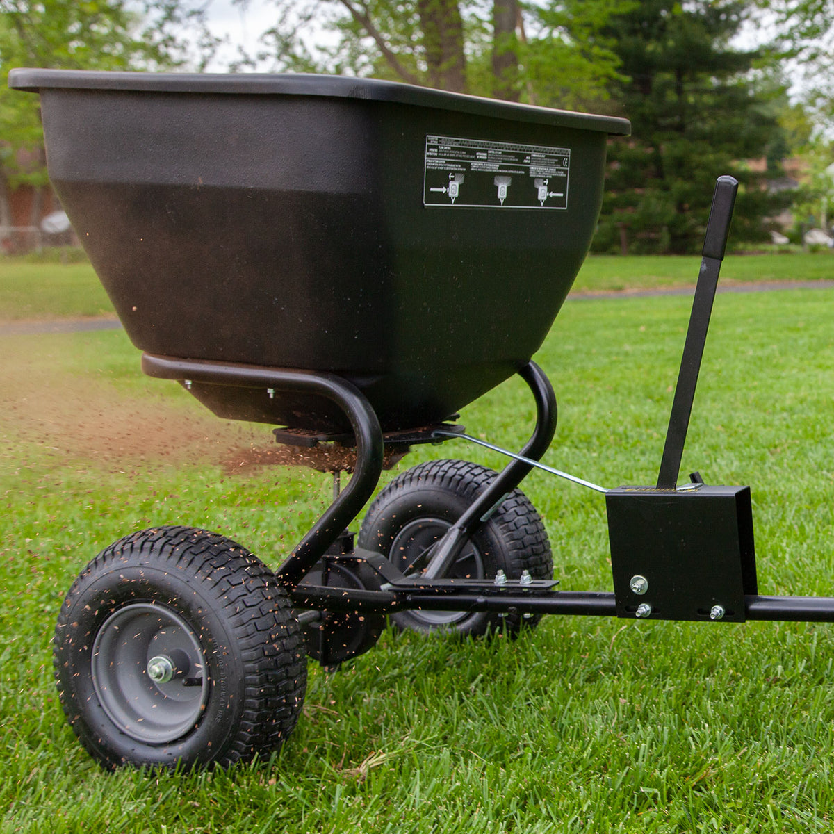 The Brinly Parts 175 Lb. Tow-Behind Spreader | BS36BH, featuring large tires, a handle, control panel, and universal hitch, effortlessly spreads materials over lawns. Its rust-proof polyethylene construction ensures durability against a backdrop of trees and grass.