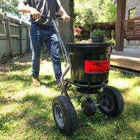 A person in jeans and a gray shirt pushes a black Brinly Parts 50 LB. Push Spreader with Deflector Kit, model P20-500BHDF, across a grassy yard near a wooden porch, with a wooden fence and trees in the background.