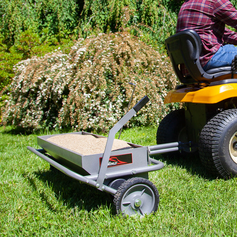 A yellow lawn tractor pulls a metal trailer loaded with sand on the grass, ready for a spike aeration task using the 40 Combination Aerator Spreader in Hammered Gunmetal (AS2-40BH-S) by Brinly Parts. White-flowered bushes frame the scene as the driver in a red plaid shirt and jeans navigates.