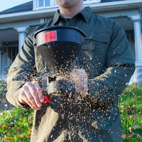 Wearing a green jacket, a person uses the Brinly Parts 5lb Capacity Handheld Spreader (HHS3-5BH) to distribute seeds or fertilizer in the yard, sending granules into the air. A house with a porch appears in the background, capturing this serene moment of garden care.