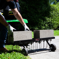 On a sunny day, someone places concrete blocks on a black metal frame with wheels, turning it into the Brinly Parts 40″ Tow-Behind Dethatcher (DT2-40BH-G). The setup attaches to a green lawn tractor amid green shrubs and a concrete path in the background.