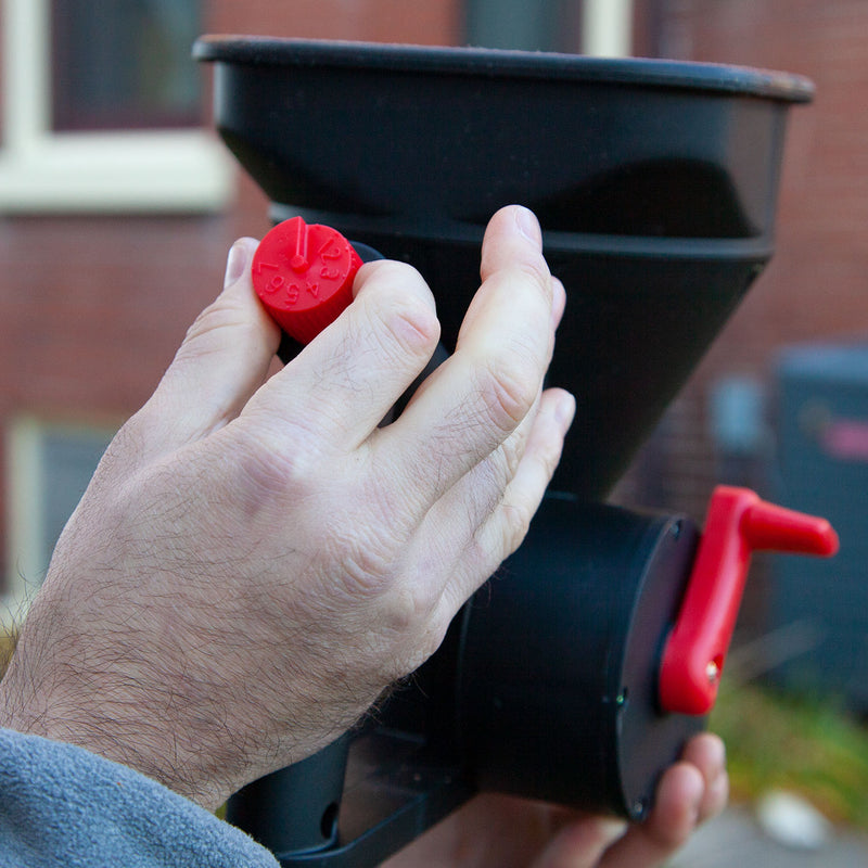 A person holds a Brinly Parts HHS3-5BH 5lb Capacity Handheld Spreader, resembling a handheld broadcast spreader. One hand adjusts the red dial while the other supports it, with part of a brick building and window visible in the background.