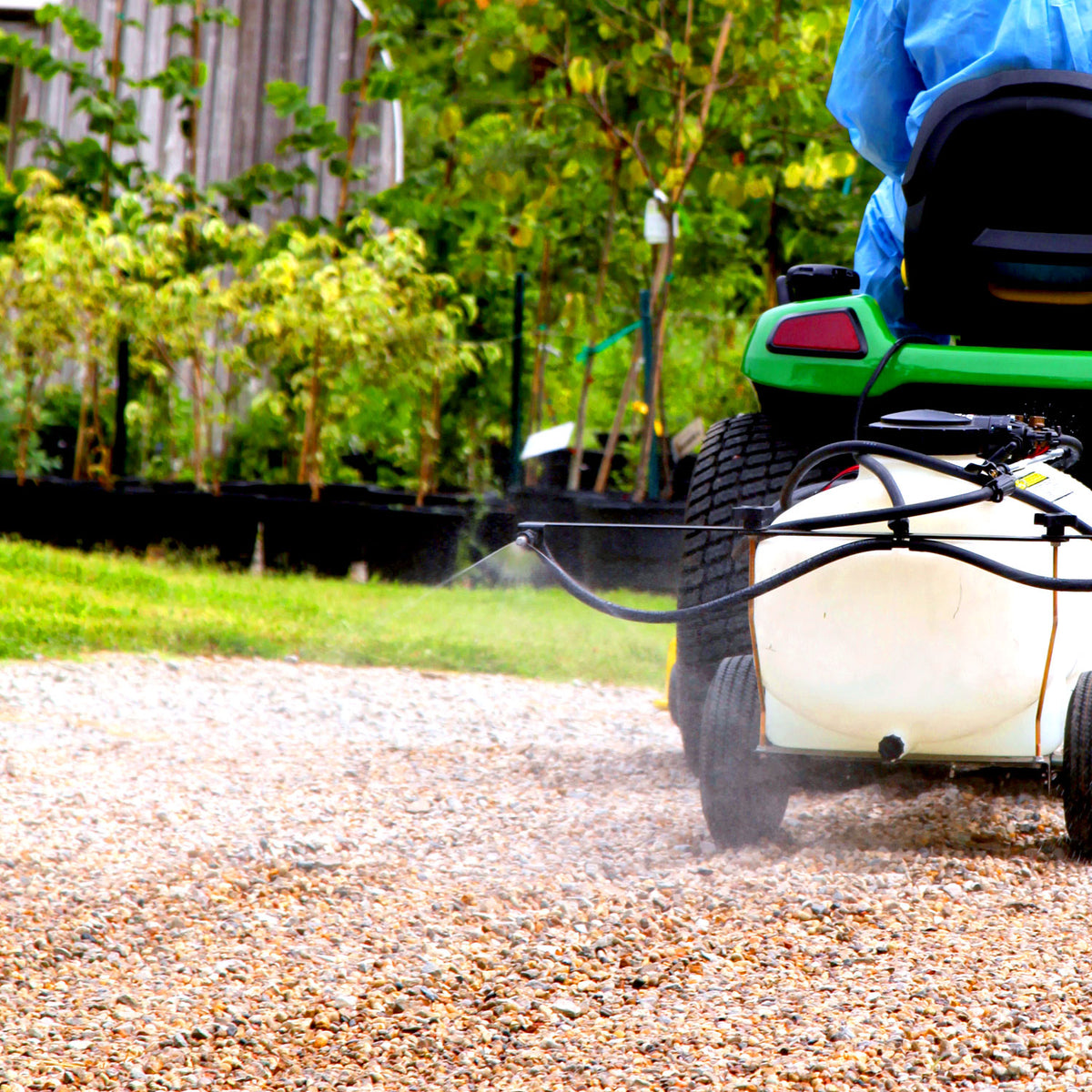 A person in a blue shirt operates a green tractor, using a Brinly Parts 25 Gallon Tow-Behind Sprayer | ST-251BH with Ultra Lo-Drift™ Spray Tips to apply liquid fertilizer on a gravel path amid greenery.