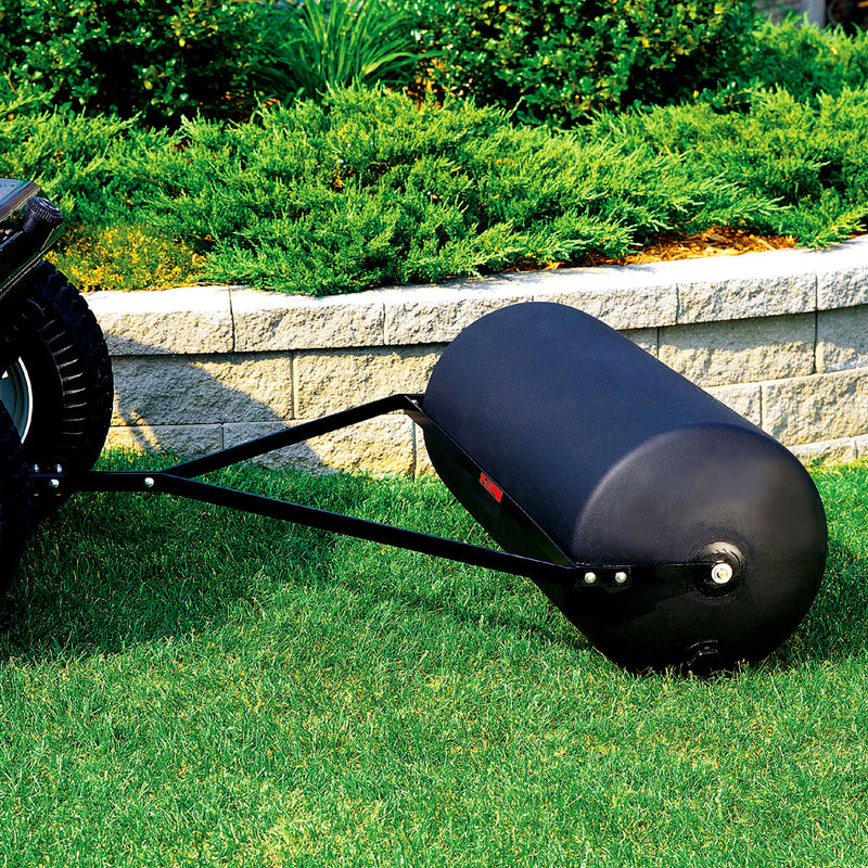 A Brinly Parts 42 Gallon Tow-Behind Poly Roller (PRT-362SBH) is hitched to a lawn tractor on lush grass. Surrounded by bushes and a stone wall, this black roller ensures a manicured lawn with its 390 lb. capacity.