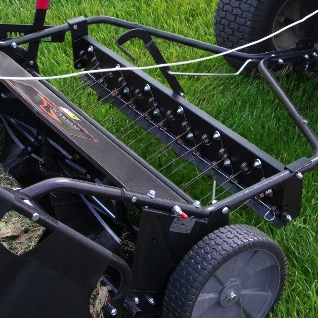 A close-up of the Brinly Parts 42″ Combo Lawn Sweeper with Dethatcher (LSDT2-42BH-G) shows metal tines and large wheels on green grass—this 2-in-1 tool efficiently removes thatch and lawn debris.