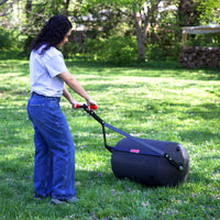 Wearing a gray shirt and blue jeans, someone uses the Brinly Parts 28 Gallon Push or Tow Poly Lawn Roller (PRC-243BH) to smooth a grassy yard, while tall trees form a scenic backdrop.