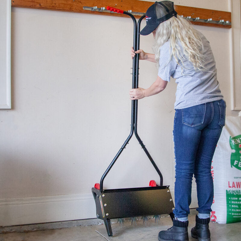 Clad in a cap, gray shirt, and jeans, the individual expertly mounts a black garden roller with red wheels using a wall-mounted tool. A Brinly Parts 18 Push Spike Aerator with 3D galvanized steel tines stands nearby, while lawn food rests on the garage floor.