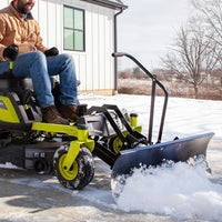 A person in a tan jacket and jeans uses a green snow plow with a Brinly Parts Front-Mount Blade for RYOBI Electric ZTRs (FB-42RYZT) attached to a riding lawn mower, clearing snow from the driveway. A house and leafless trees are visible in the background on this crisp winter day.