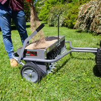 A person uses a Brinly Parts 40 Combination Aerator Spreader (AS2-40BH-S) in Hammered Gunmetal to aerate a grassy area, adding sand or seed into its container. The aerator has spiked wheels and a crank handle, with trees and shrubs visible in the background.