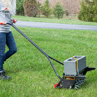 Wearing jeans and a gray shirt, a person expertly maneuvers the Brinly Parts 18 Push Spike Aerator with 3D Galvanized Steel Tines, using a concrete block for extra weight, as they diligently work across the grassy lawn against a backdrop of trees.