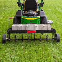 A Brinly Parts 48 Tow-Behind Plug Aerator (PA-482BH), featuring sturdy steel tines, is attached to a green lawn tractor on the grass. The aerator is loaded with cinder blocks for added weight, enhancing its effectiveness in penetrating deep into the root zone.