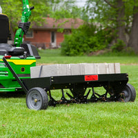 A green lawn tractor tows the Brinly Parts 48″ Tow-Behind Plug Aerator (PA-482BH) with steel tines, loaded with concrete blocks for added weight. It efficiently aerates the grass, while a brick house and lush trees complete the scenic view.