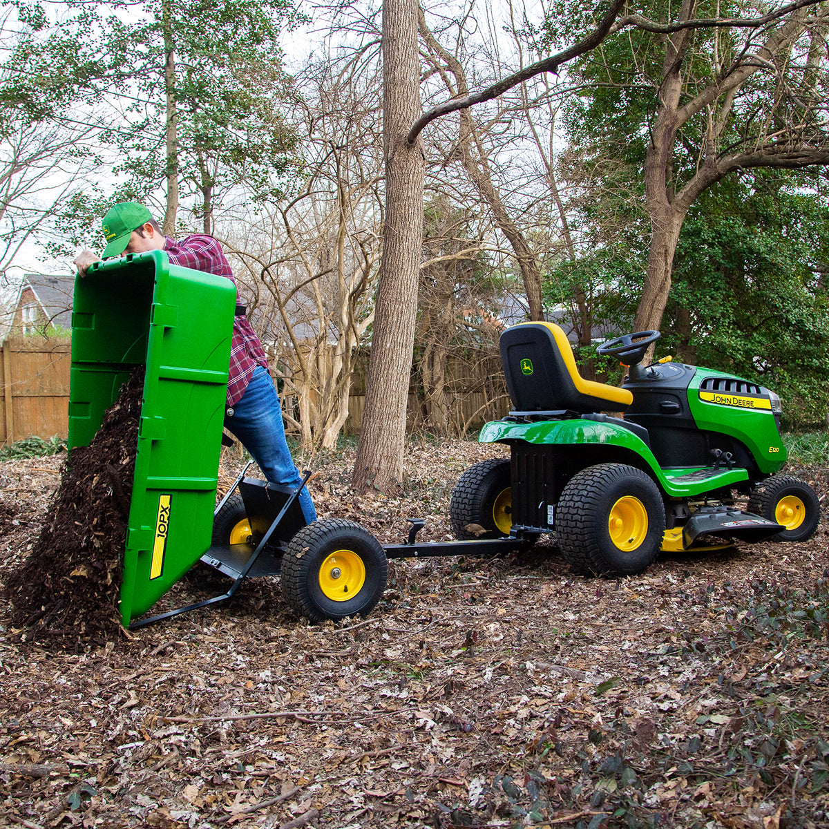 Wearing a green cap and plaid shirt, a person expertly operates a John Deere lawn tractor with the 180 Degree Full Dump Cart, effortlessly unloading leaves in a wooded area at its full 180-degree dump angle, demonstrating its impressive 650 lb. hauling capacity.