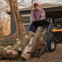 A person in a plaid shirt unloads large logs from a Brinly Parts 10 Cu. Ft. Poly Cart (PCT-101BH) next to a shed and trees in a yard.