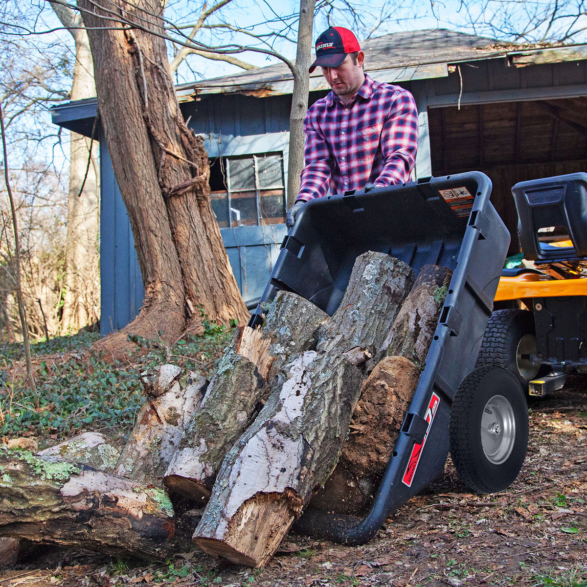 A man in a red cap and plaid shirt unloads logs from a Brinly Parts 10 Cu. Ft. Poly Cart | PCT-101BH, hitched to a yellow lawn tractor beside a shed in a wooded backyard.