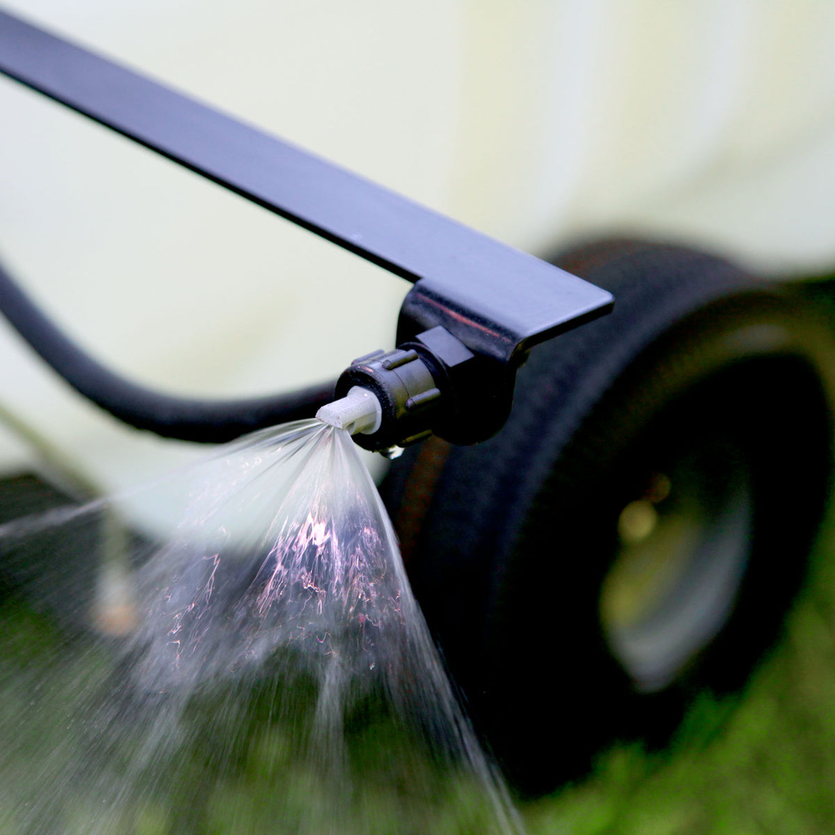 Close-up of a black nozzle from the Brinly Parts 25 Gallon Tow-Behind Sprayer (ST-251BH) releasing a fine mist. The nozzle is on a horizontal bar, with a blurred background featuring a black tire on grass.