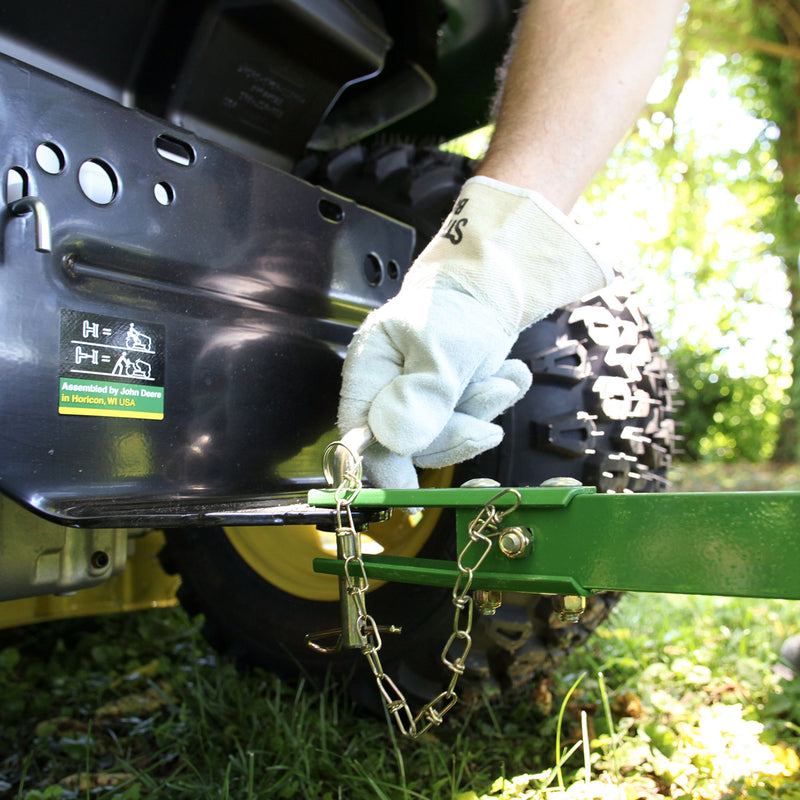 A person wearing a glove adjusts a green lever attached to a tethered hitch pin on a John Deere 36 Lawn Roller (PRT-361SJD/LPPRT36JD) with grassy ground and a wheel visible outdoors amidst distant trees.