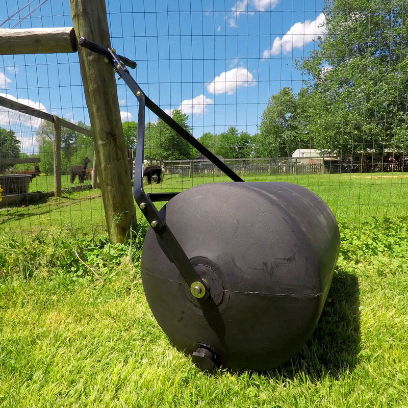 A Brinly Parts 28 Gallon Push or Tow Poly Lawn Roller (PRC-242BH) sits on green grass next to a wire fence, under a blue sky with scattered clouds, surrounded by trees and buildings.