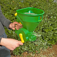 A person uses the John Deere Handheld Spreader LP35445-A to distribute seeds. The ergonomic trigger handle, adjustable flow gate, and yellow design make it efficient. Green foliage flourishes in the background as seeds spread evenly.